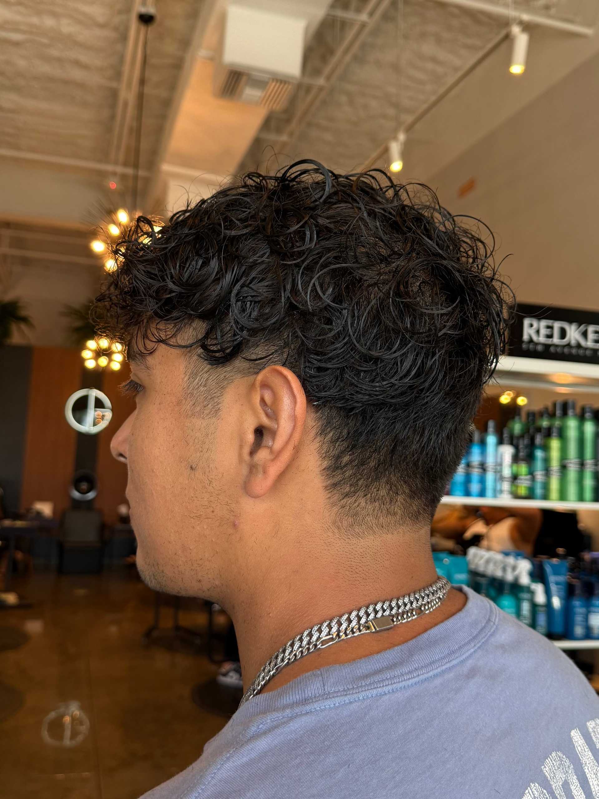 Man with curly hairstyle in a salon, wearing a chain necklace and a light blue shirt.